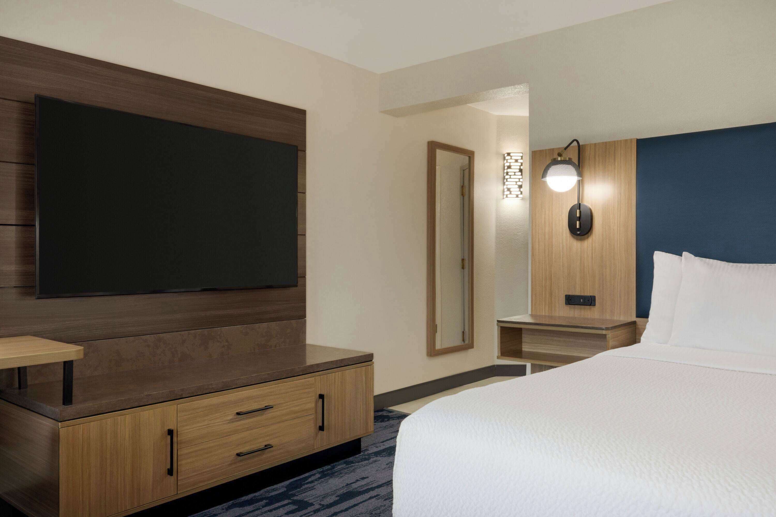 Habitación de hotel con una cama grande, un televisor de pantalla plana montado en la pared, un aparador de madera con cajones, una mesita de noche, un espejo y una lámpara de pared.
