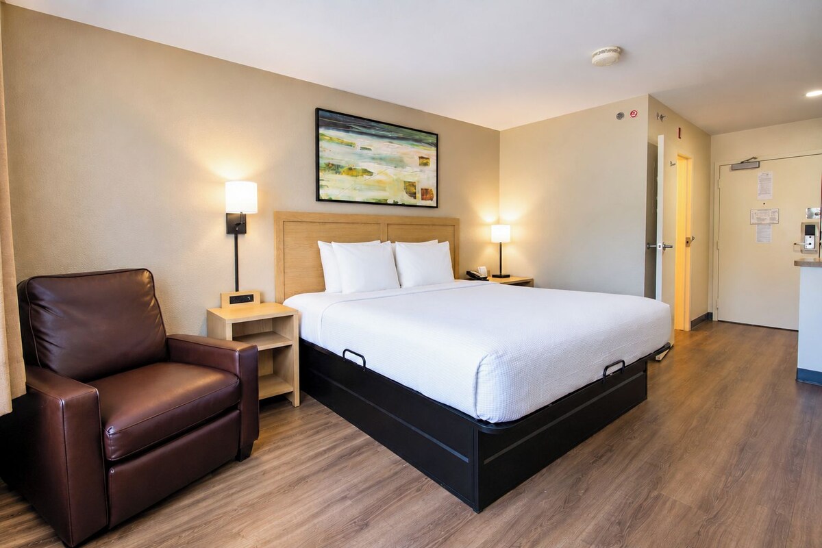 A hotel room with a bed, a chair, a nightstand, a lamp, and a painting on the wall.