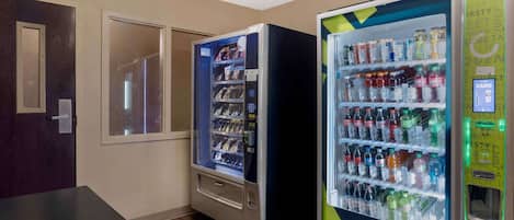 A room with two vending machines stocked with beverages and snacks, a wooden floor, and a door with a frosted glass panel.