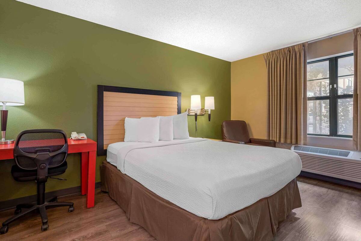 Een hotelkamer met een bed, bureau, stoel, lamp en een airconditioning.