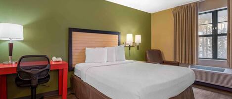 A hotel room with a bed, desk, chair, lamp, and air conditioning unit.