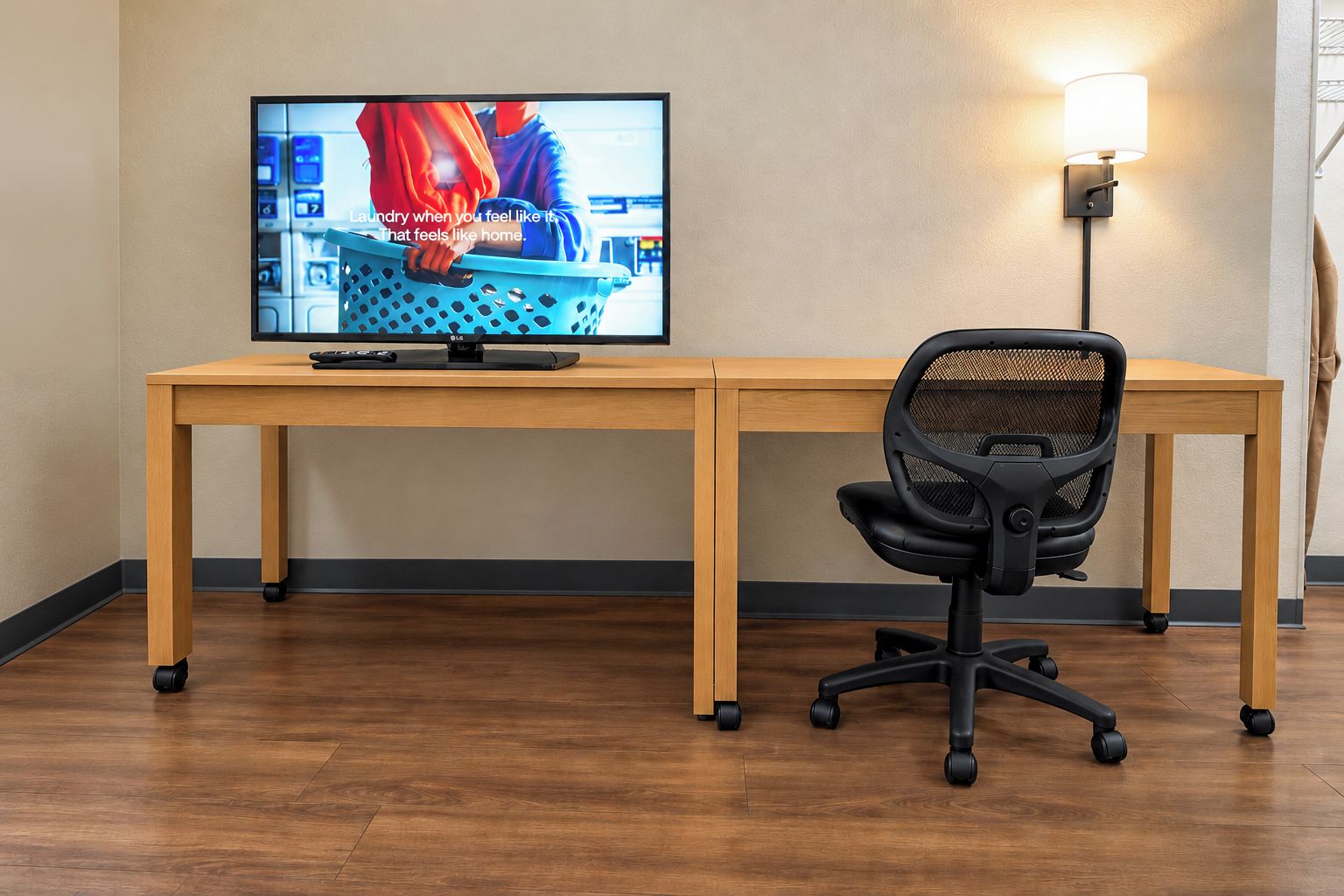 A room with a wooden desk, a black office chair, a television, and a wall-mounted lamp.