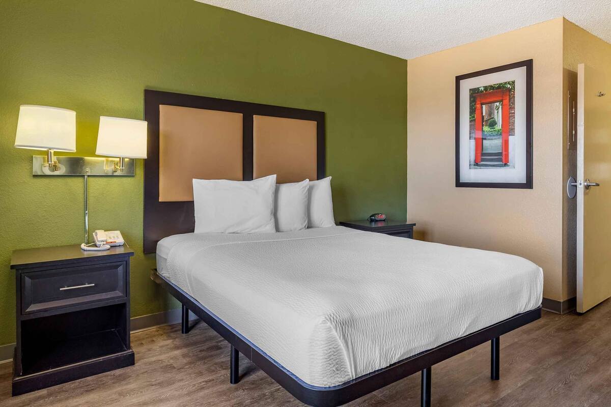 A neatly made bed with a white comforter and pillows, a bedside table with a lamp, and a framed picture on the wall.