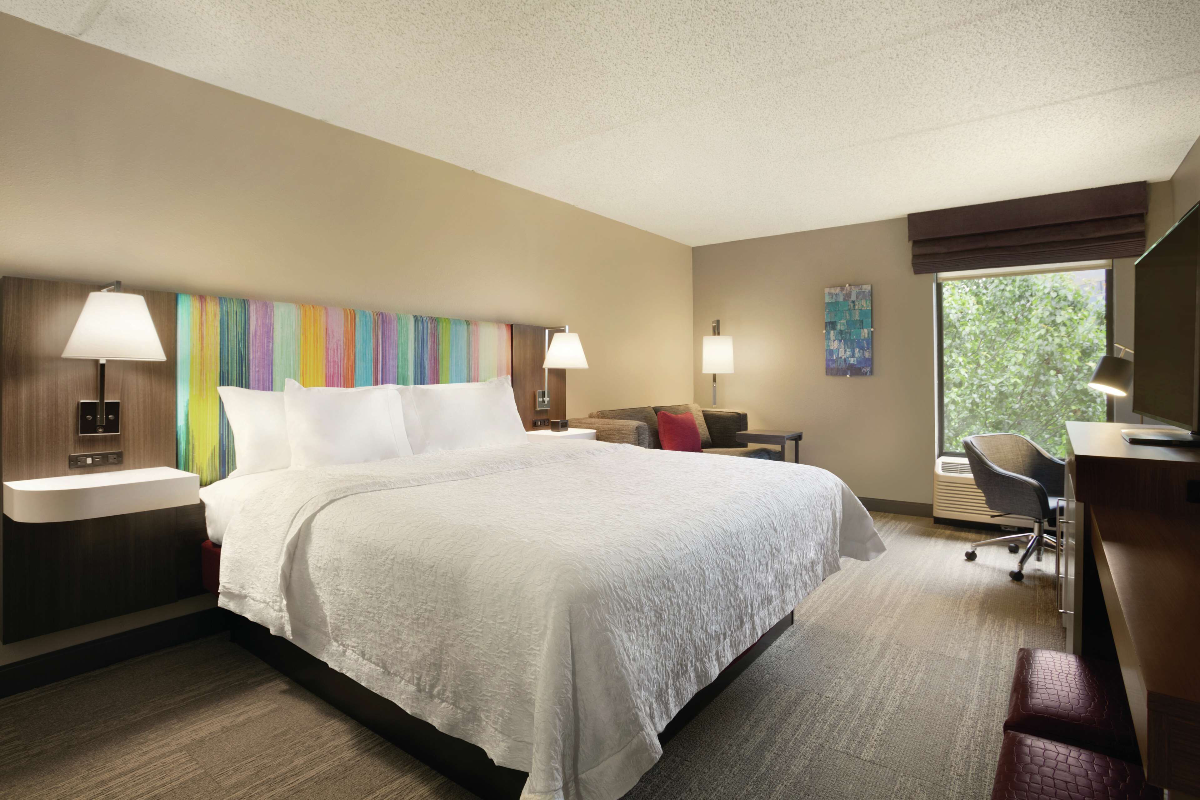 A hotel room with a large bed, bedside tables with lamps, a desk with a chair, and a window with a view of greenery.