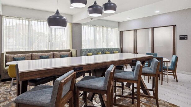 A modern hotel dining area with a long wooden table, six upholstered chairs, and three pendant lights. - Hampton Inn Houston-Brookhollow (Houston)