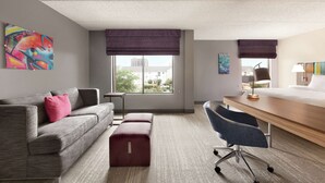 A modern hotel room with a grey sofa, a wooden desk, a blue office chair, and a window with a view of buildings. - Hampton Inn Houston-Brookhollow (Houston)