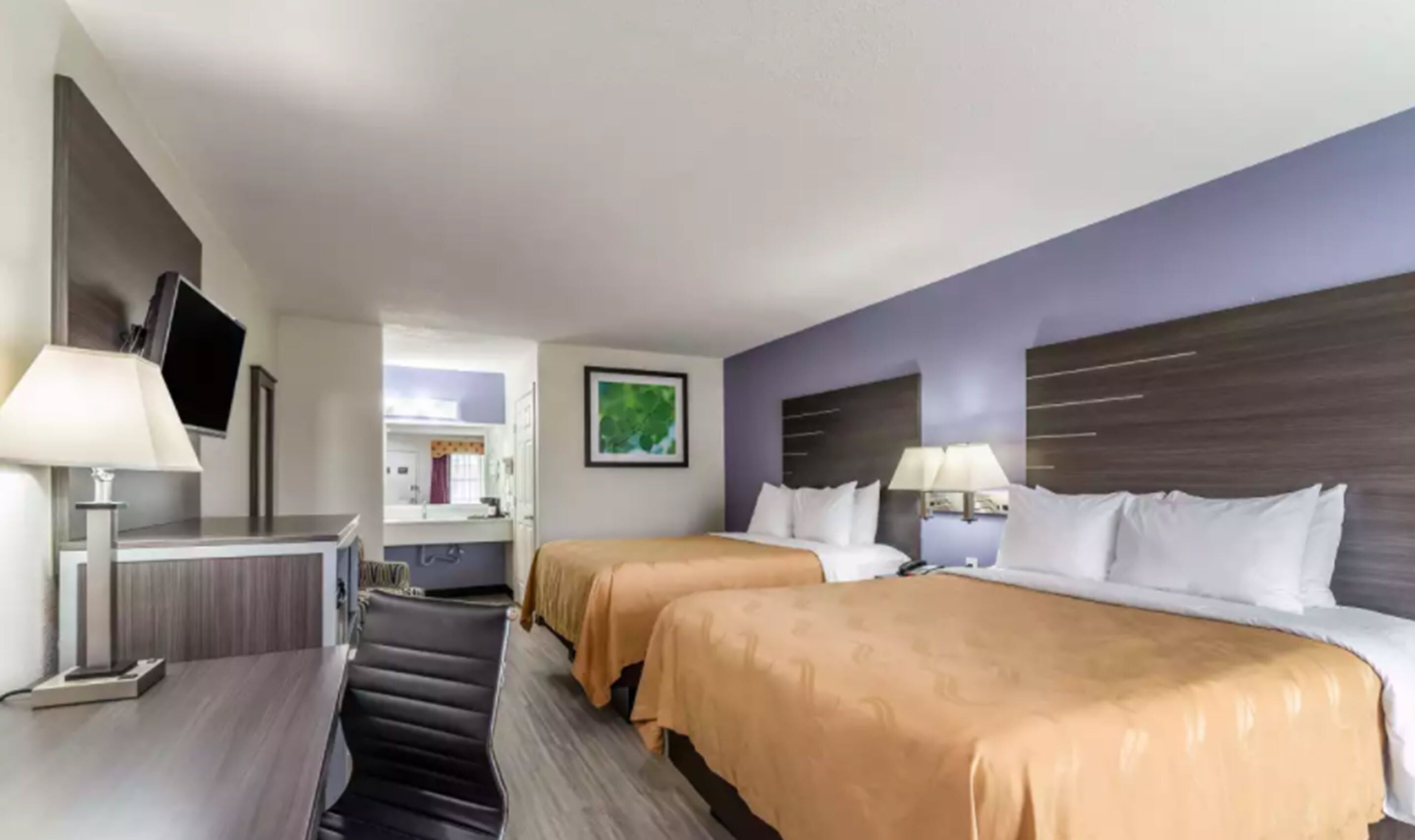 Habitación de hotel con dos camas, un escritorio, televisión y baño visible a través de una puerta abierta.