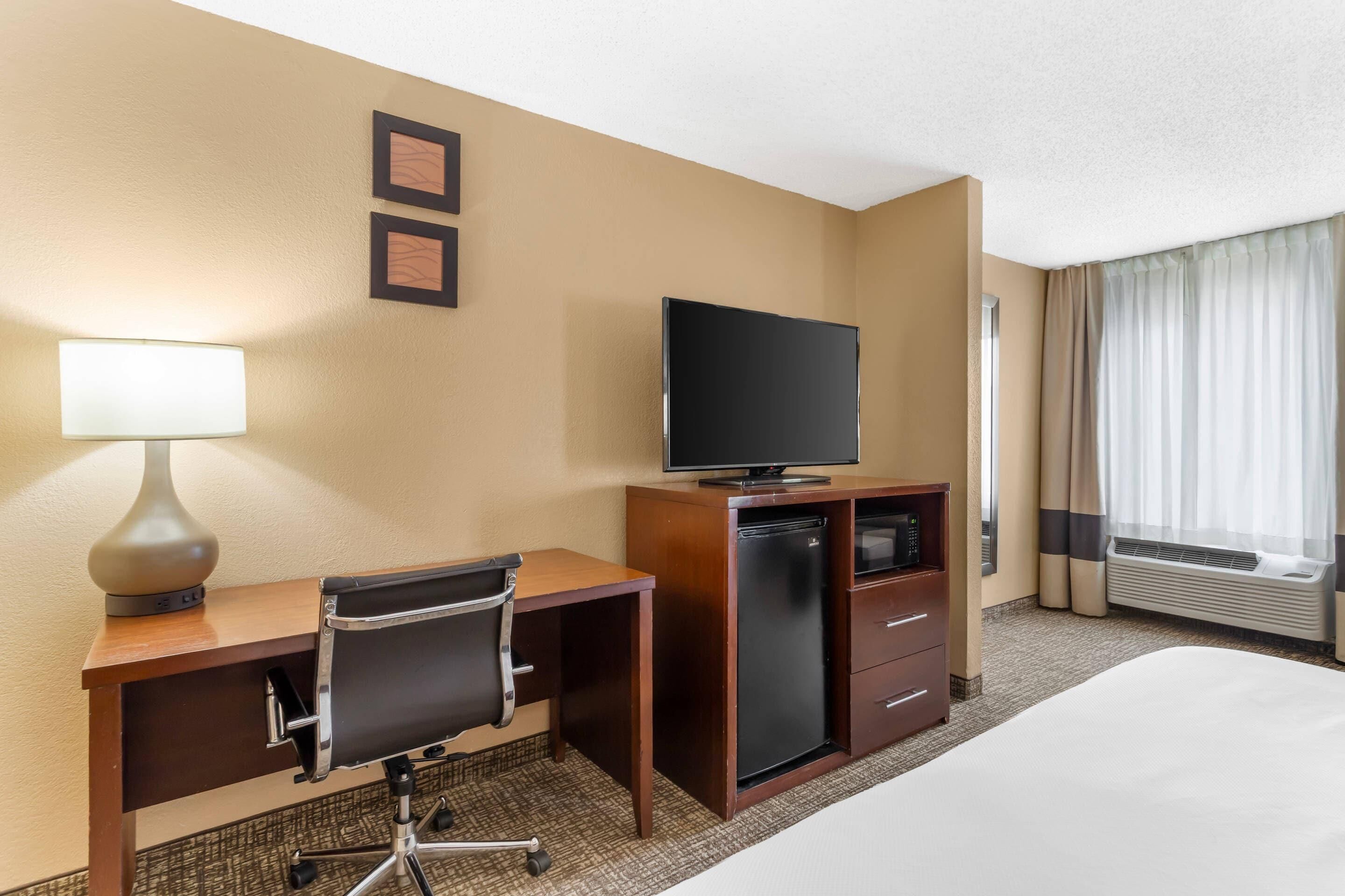 A hotel room with a bed, a desk with a chair, a television, a mini-fridge, a lamp, and a window with curtains.