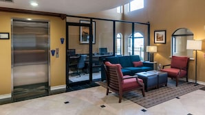 A lobby with a modern elevator, a seating area with a blue sofa and red chairs, and a glass partition separating the lobby from the office space. - Comfort Inn North Conroe (Conroe)