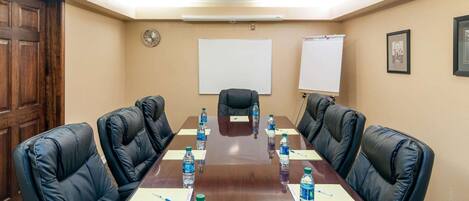 Una sala de conferencias con una larga mesa, sillas de cuero negro, botellas de agua, tarjetas de identificación, un reloj, una pizarra y un cuadro en la pared.