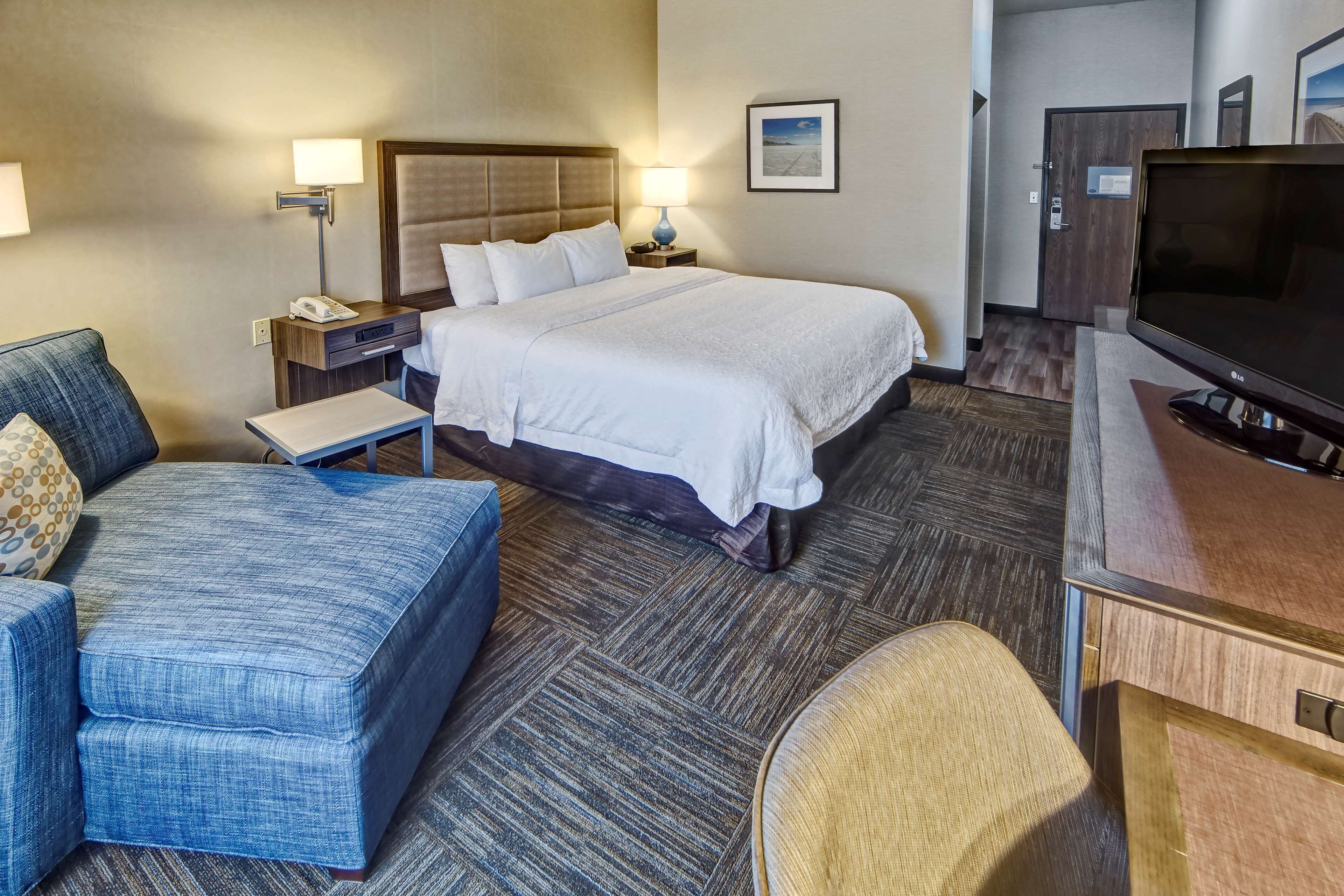 Habitación de hotel con una cama grande, un escritorio con televisor, una zona de estar con una silla y reposapiés, y una mesita de noche con lámpara y teléfono.