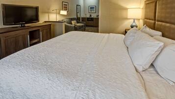 A hotel room with a large bed, a flat-screen TV, a desk with a chair, a kitchenette, and a bathroom visible in the background.