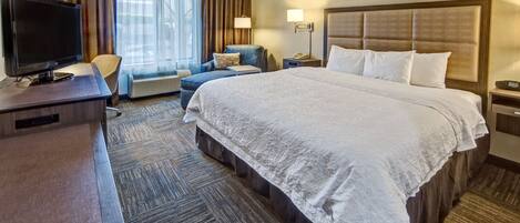 A hotel room with a large bed, a desk with a television, a chair, a sofa, and a window with curtains.