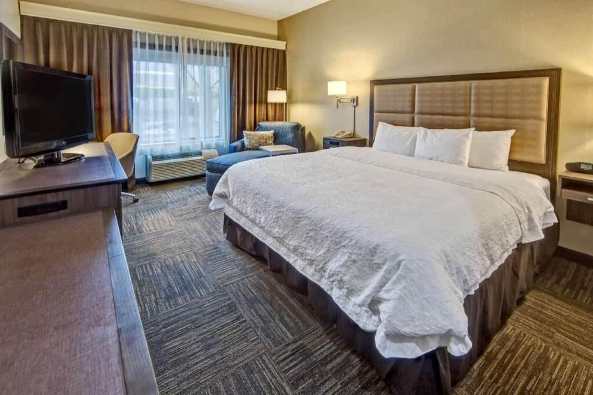 A hotel room with a large bed, a desk with a TV, a chair, a lamp, and a window with curtains.