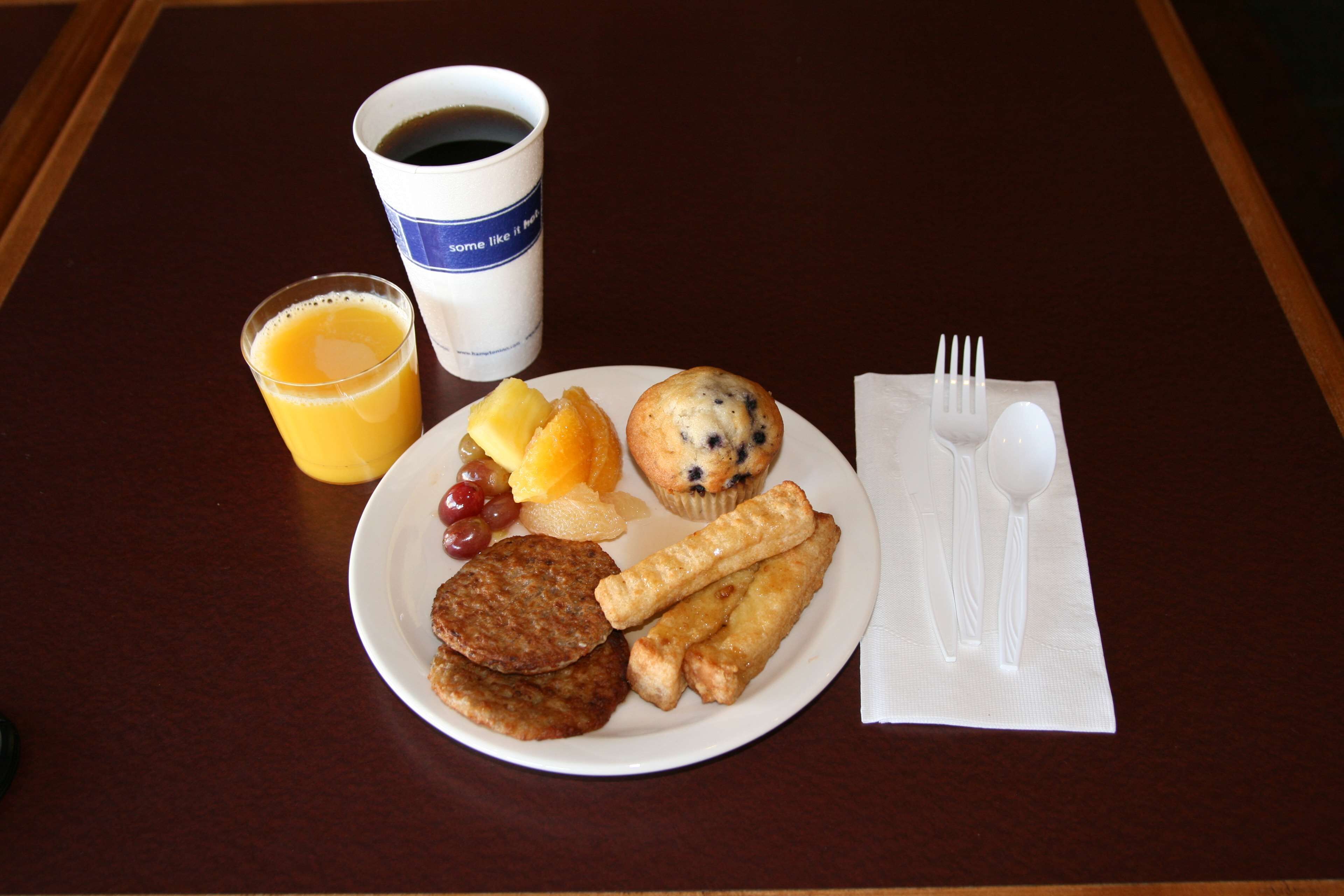 A breakfast plate with pancakes, a muffin, sausage links, and a glass of orange juice on a table.