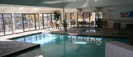 Piscina interna com paredes de vidro transparente, piso ladrilhado e várias áreas de estar com cadeiras e mesas.