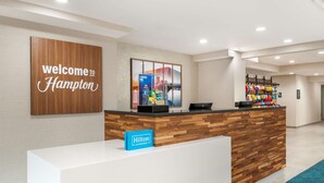Un mostrador de recepción con un cartel que dice 'Welcome to Hampton', un cartel de 'Hilton Honors' y una presentación de artículos de tocador.