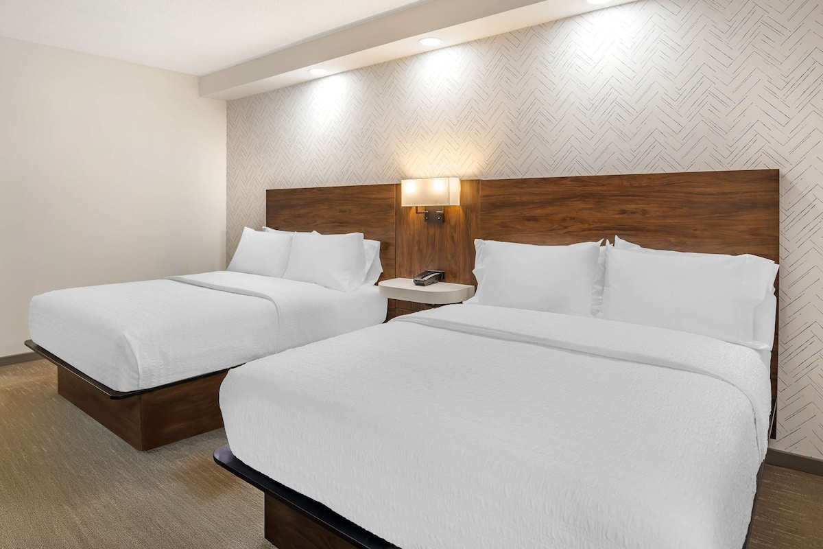 Habitación de hotel con dos camas, cabeceros de madera, una mesita de noche con lámpara y pared con motivo de espiga.
