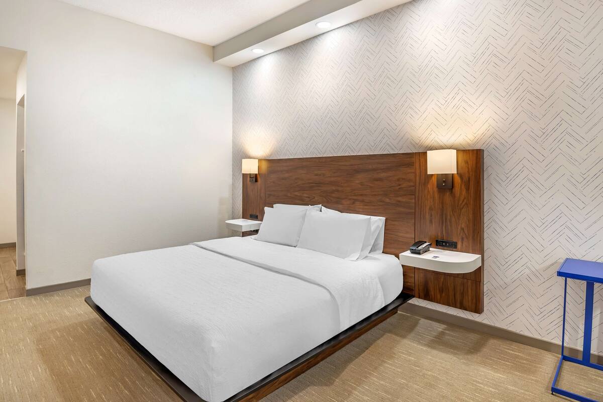 Una habitación de hotel moderna con una cama grande, cabecera de madera, dos mesitas de noche con lámparas y una mesita auxiliar azul.