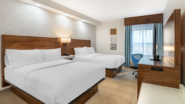 Habitación de hotel con dos camas, un escritorio, una silla y un amplio ventanal con cortinas.