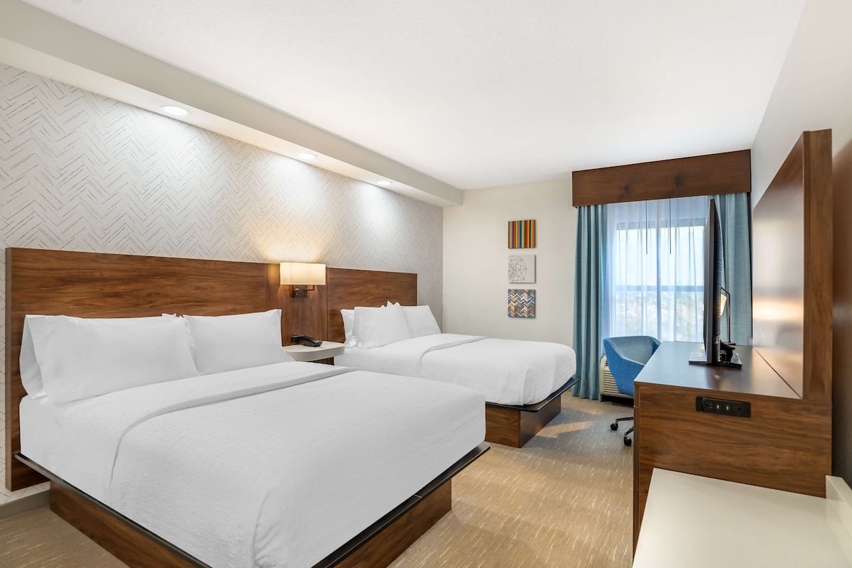 Habitación de hotel con dos camas, un escritorio, una silla y un amplio ventanal con cortinas.