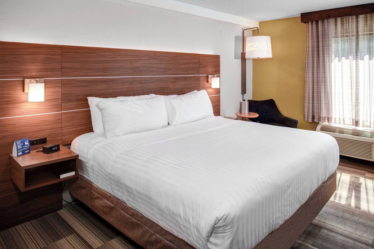 A hotel room with a large bed, wooden headboard, bedside tables, a nightstand, a lamp, a chair, and a window with curtains.