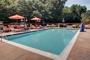 A rectangular swimming pool with clear blue water, surrounded by lounge chairs and umbrellas, set against a backdrop of trees.