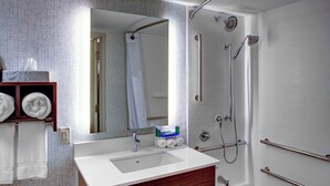 Ein modernes Badezimmer mit einem weißen Waschbecken, einem Spiegel und einer Dusche. Handtücher liegen auf einem Regal und ein Toilettenpapierhalter ist an der Wand angebracht.
