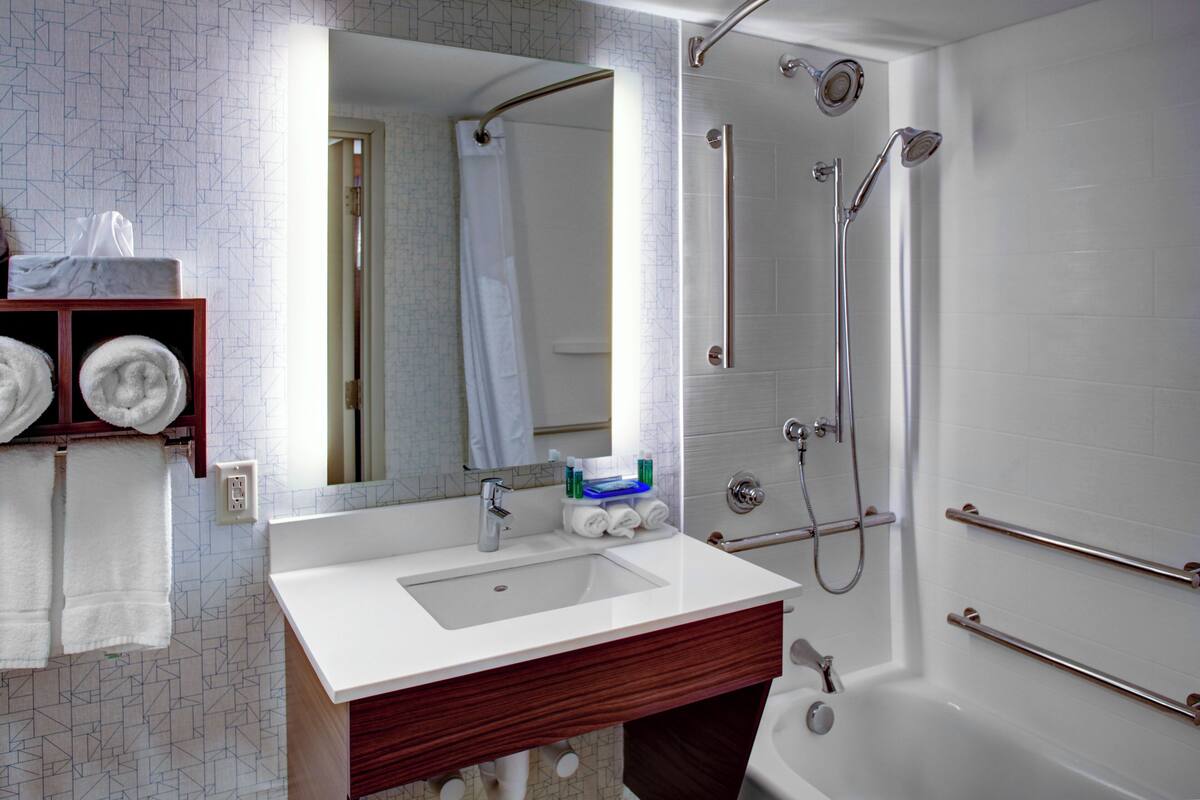 A modern bathroom with a white sink, mirror, and shower. There are towels on a shelf and a toilet paper holder on the wall.