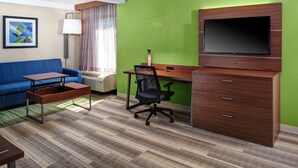 Ein Hotelzimmer mit einem blauen Sofa, einem Holztisch, einem schwarzen Bürostuhl, einem Fernseher auf einer Holzkommode und einem gerahmten abstrakten Kunstwerk an der Wand.
