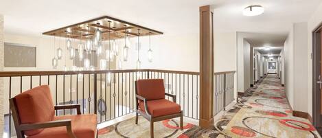 Un couloir d’hôtel avec un tapis à motifs, deux chaises en bois, une rambarde et un luminaire au plafond.