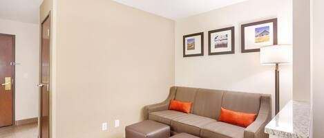 A modern hotel room with a beige wall, a brown sofa with orange cushions, a small table, and a lamp.
