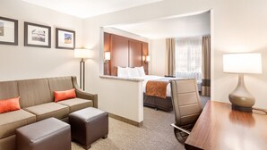Ein Hotelzimmer mit einer Couch, einem Hocker, einem Schreibtisch mit Lampe, einem Bett mit Kopfteil und gerahmten Bildern an der Wand.