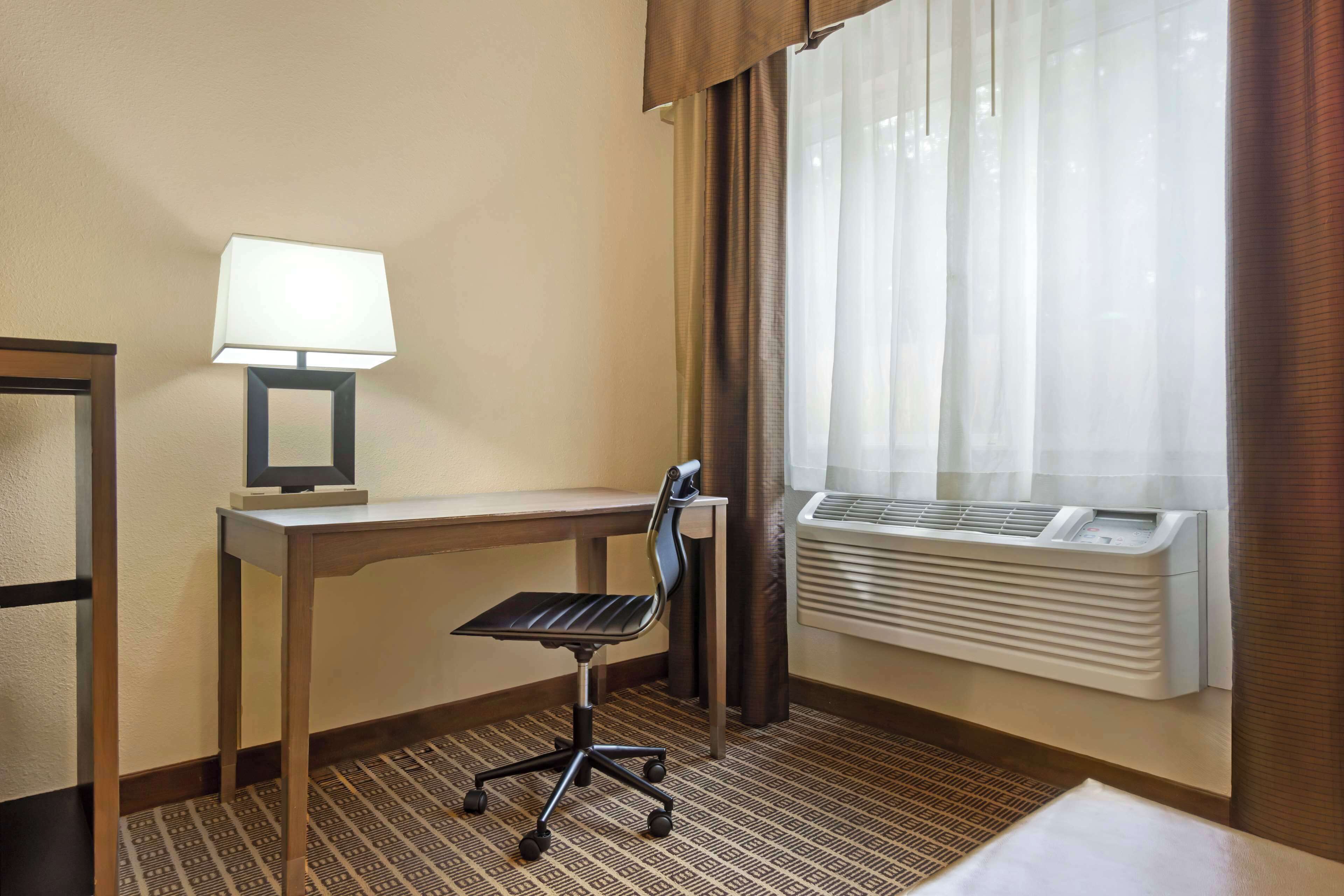 A hotel room with a desk, chair, lamp, air conditioning unit, and a window with curtains.