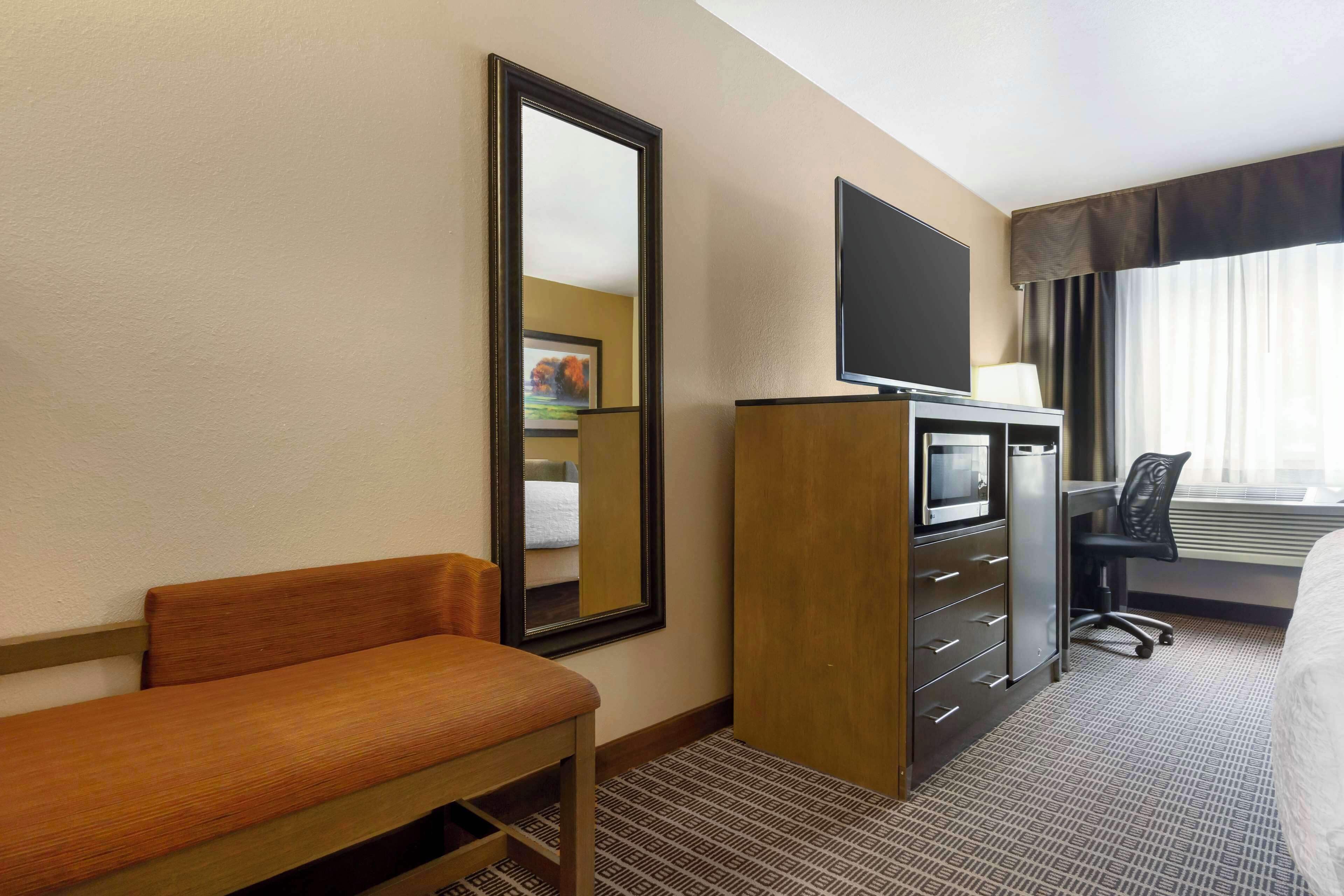 Een hotelkamer met een bed, een bureau met stoel, een televisie, een spiegel en een raam met gordijnen.
