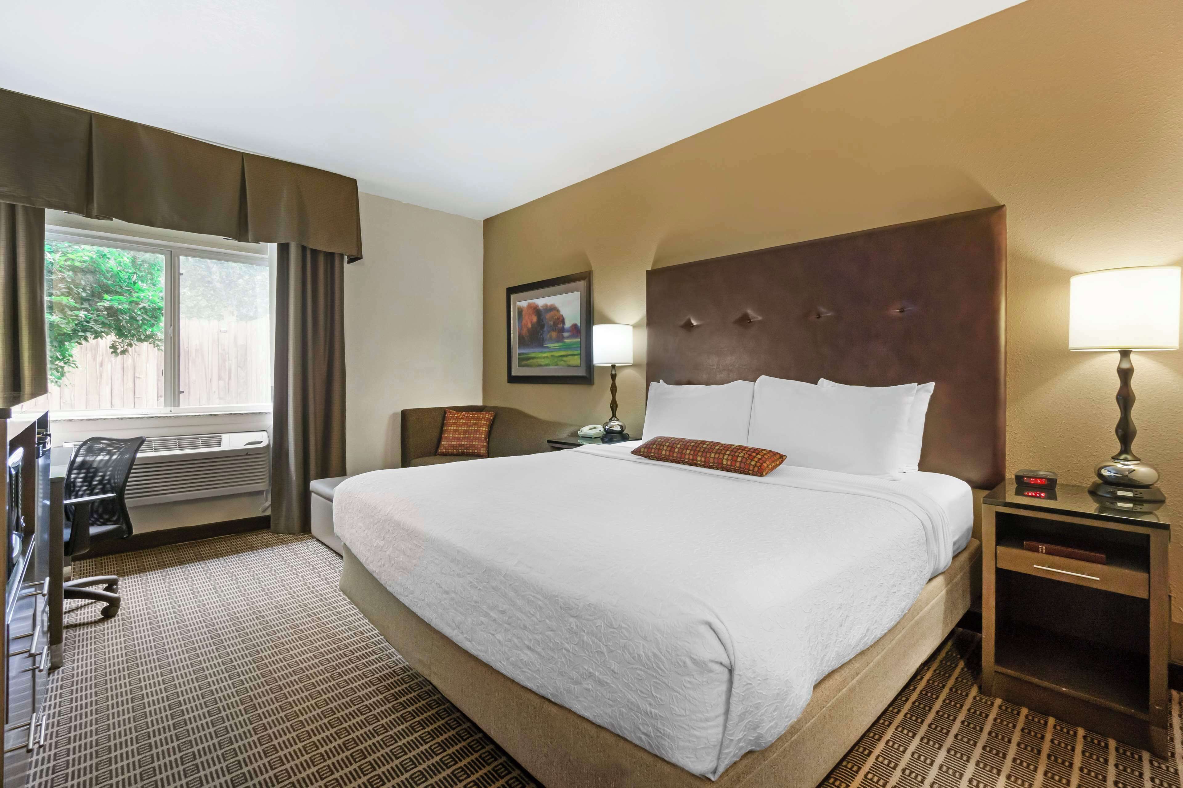 Habitación de hotel con una cama grande, un escritorio con silla, una mesita de noche con lámpara y una ventana con cortinas.