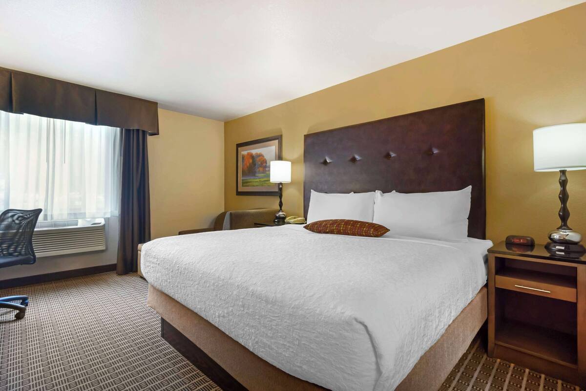 Une chambre d’hôtel avec un grand lit, un bureau avec une lampe, une chaise et une fenêtre avec des rideaux.