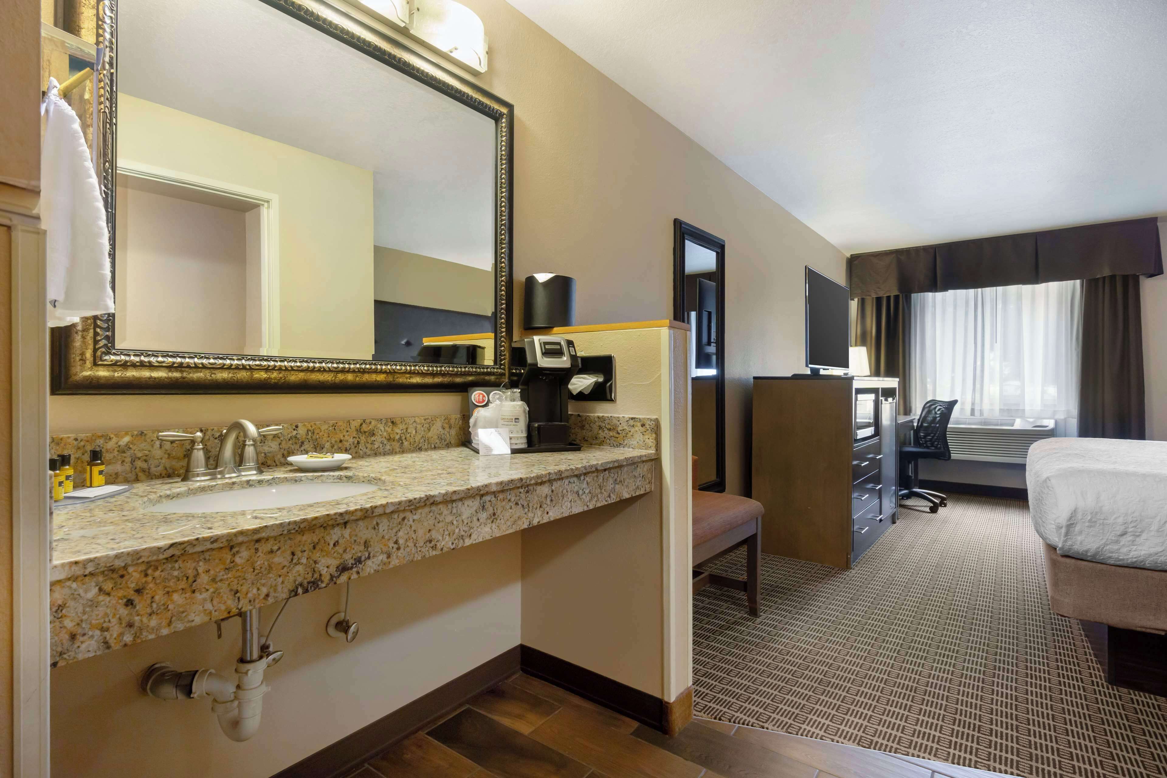 Quarto de hotel com pia de bancada de granito, um espelho grande, máquina de café, uma cadeira, cama e janela com cortinas.