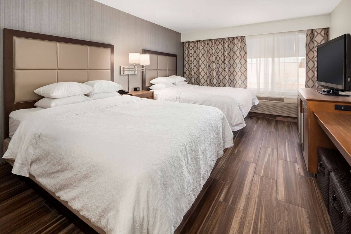 A hotel room with two beds, a desk with a television, a bedside table with lamps, and a patterned curtain.