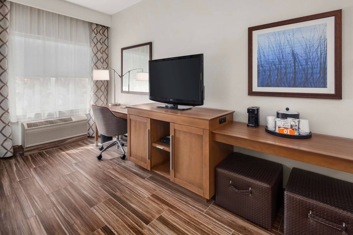 A hotel room with a wooden desk, a television, a framed picture, a coffee maker, and wicker storage bins.