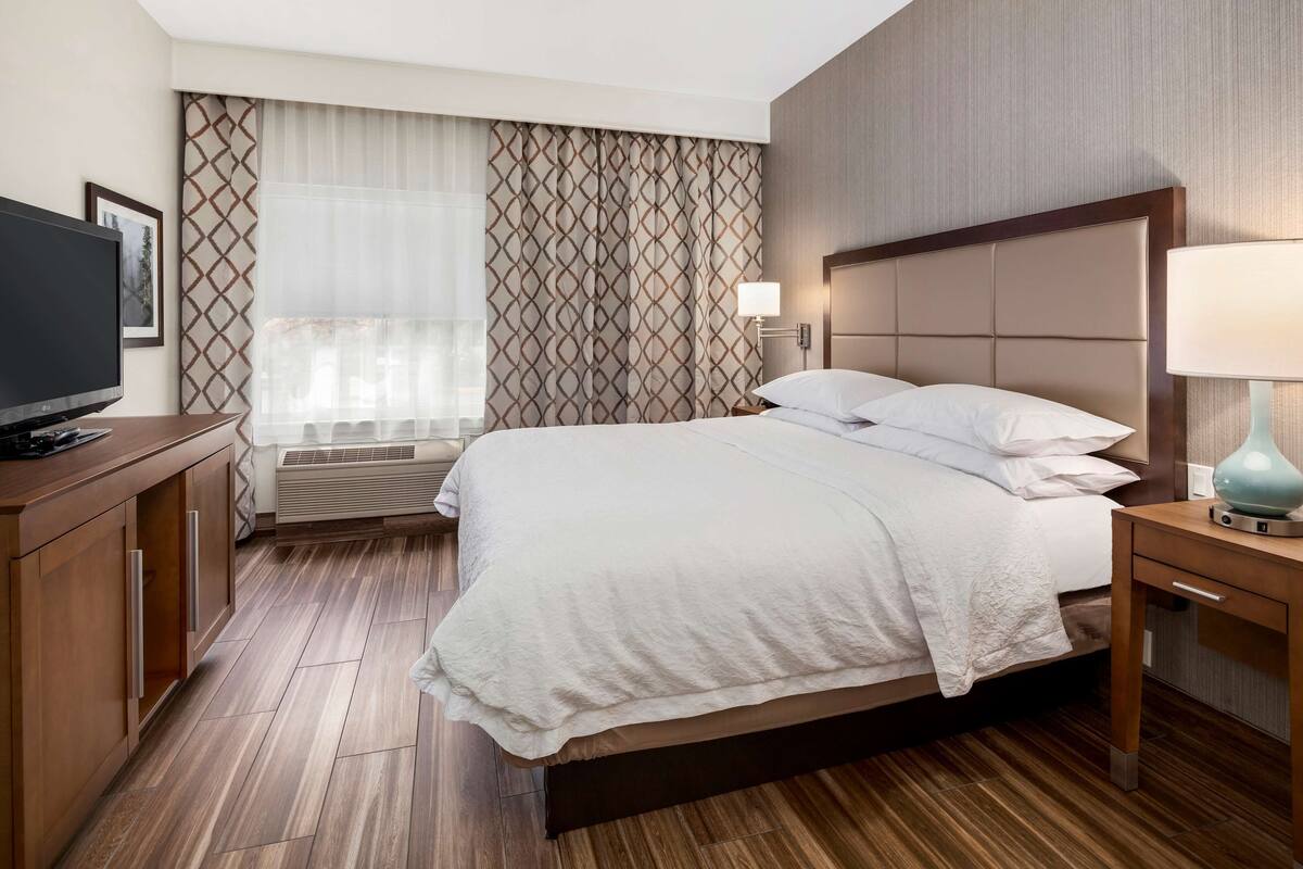 A hotel room with a large bed, a television, a desk with a lamp, and a window with curtains.
