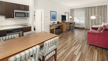 A modern hotel room with a wooden dining table, checkered chairs, a red sofa, a TV, and a small kitchenette.