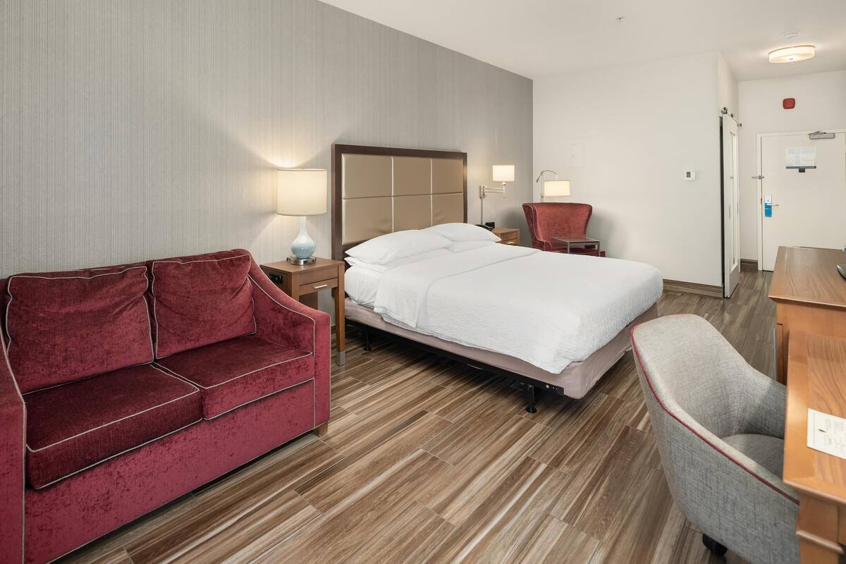 A hotel room with a bed, a red sofa, a grey armchair, a wooden desk, and a nightstand with a lamp.