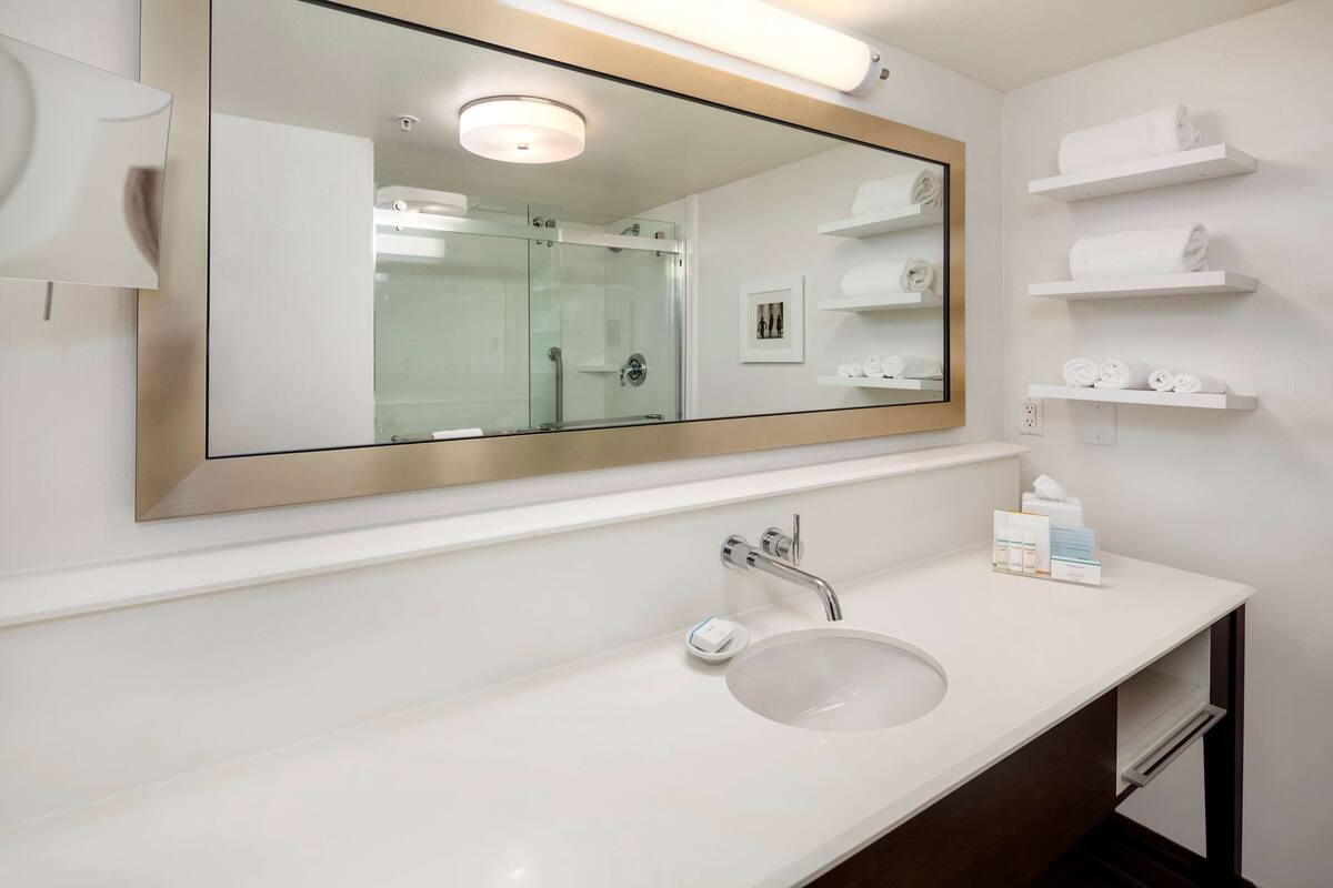 A modern bathroom with a large mirror, a white sink, and built-in shelves with towels.