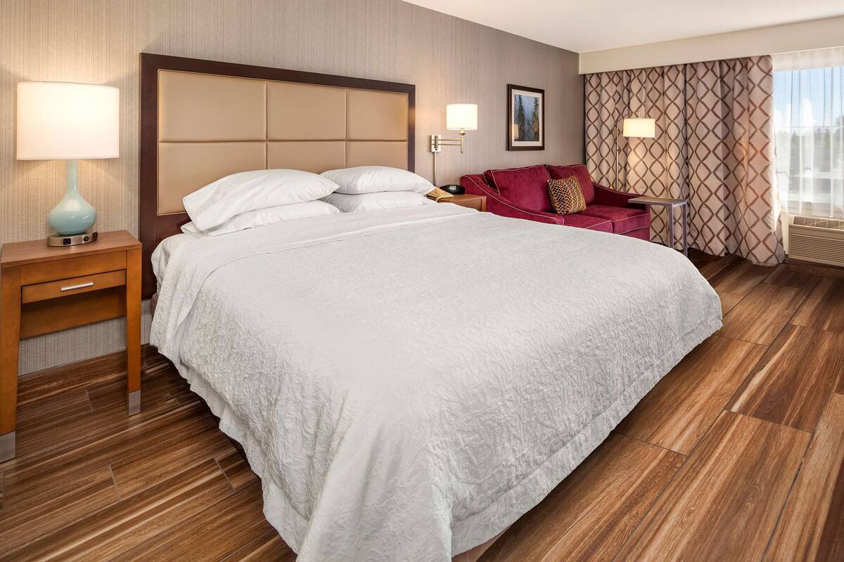 A hotel room with a large bed, a nightstand with a lamp, a red armchair, and a patterned curtain.