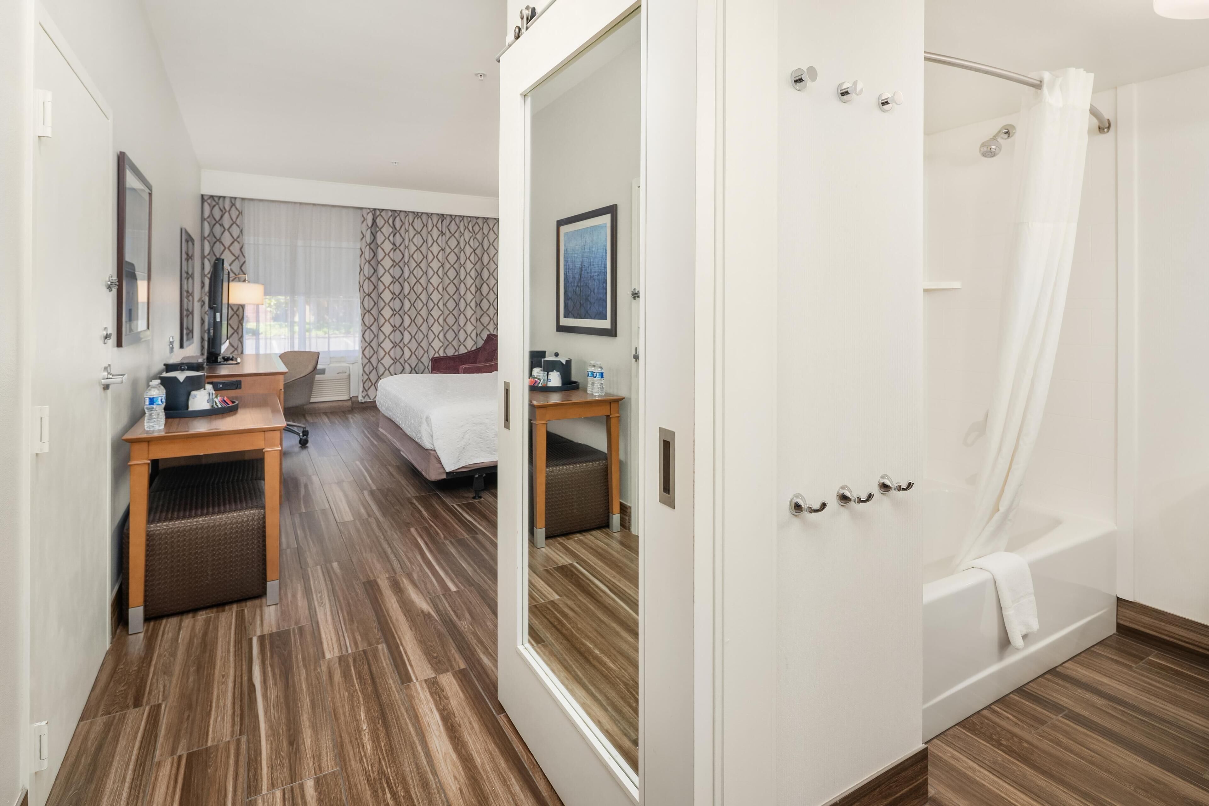 A hotel room with a wooden floor, a bed, a desk with a chair, a small table with a lamp, a shower area, and a framed picture on the wall.