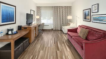Habitación de hotel con piso de madera, sofá rojo, televisor, escritorio con computadora y ventana con cortinas estampadas.