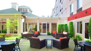 An outdoor patio area with wicker furniture and a small round table, surrounded by greenery and a building facade. - Hilton Garden Inn Seattle/Renton (Renton)