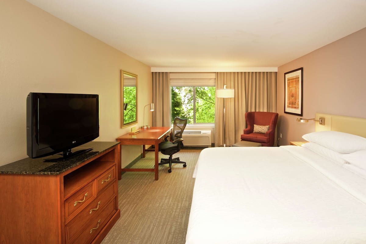 A hotel room with a bed, a desk with a TV, a chair, and a window with curtains.