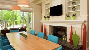 A modern dining area with a wooden table, blue chairs, a fireplace, and a TV mounted above the mantel. - Hilton Garden Inn Seattle/Renton (Renton)